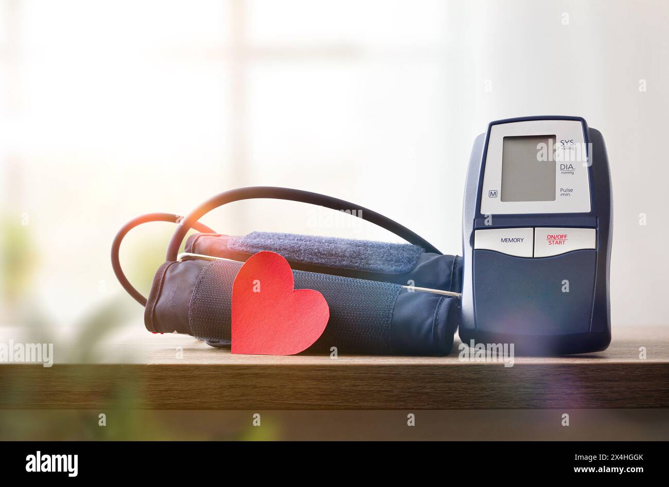 Moniteur de pression artérielle numérique bleu pour la maison sur table en bois avec découpe de coeur rouge pour la surveillance de la pression artérielle et fenêtre avec rideaux dans le BA Banque D'Images