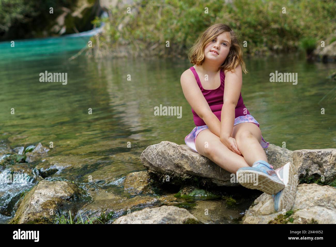 Fille de huit ans en maillot de bain rouge assise sur les rochers au bord d'une rivière cristalline souriant à la caméra Banque D'Images