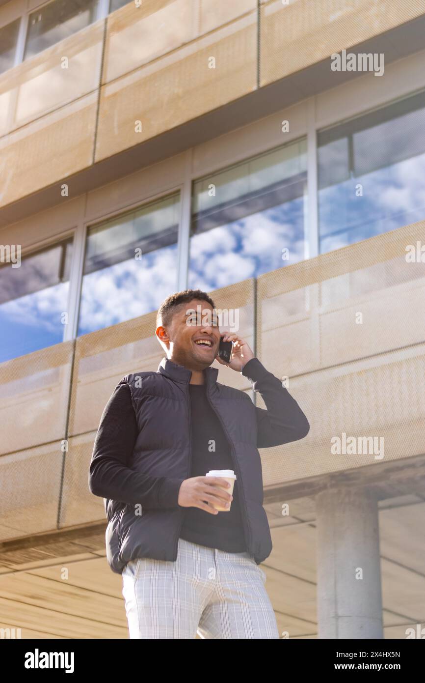 Jeune homme d'affaires décontracté intelligent marchant dans la rue parlant au téléphone tenant un verre de café. Gars souriant en veste parlant au téléphone Banque D'Images