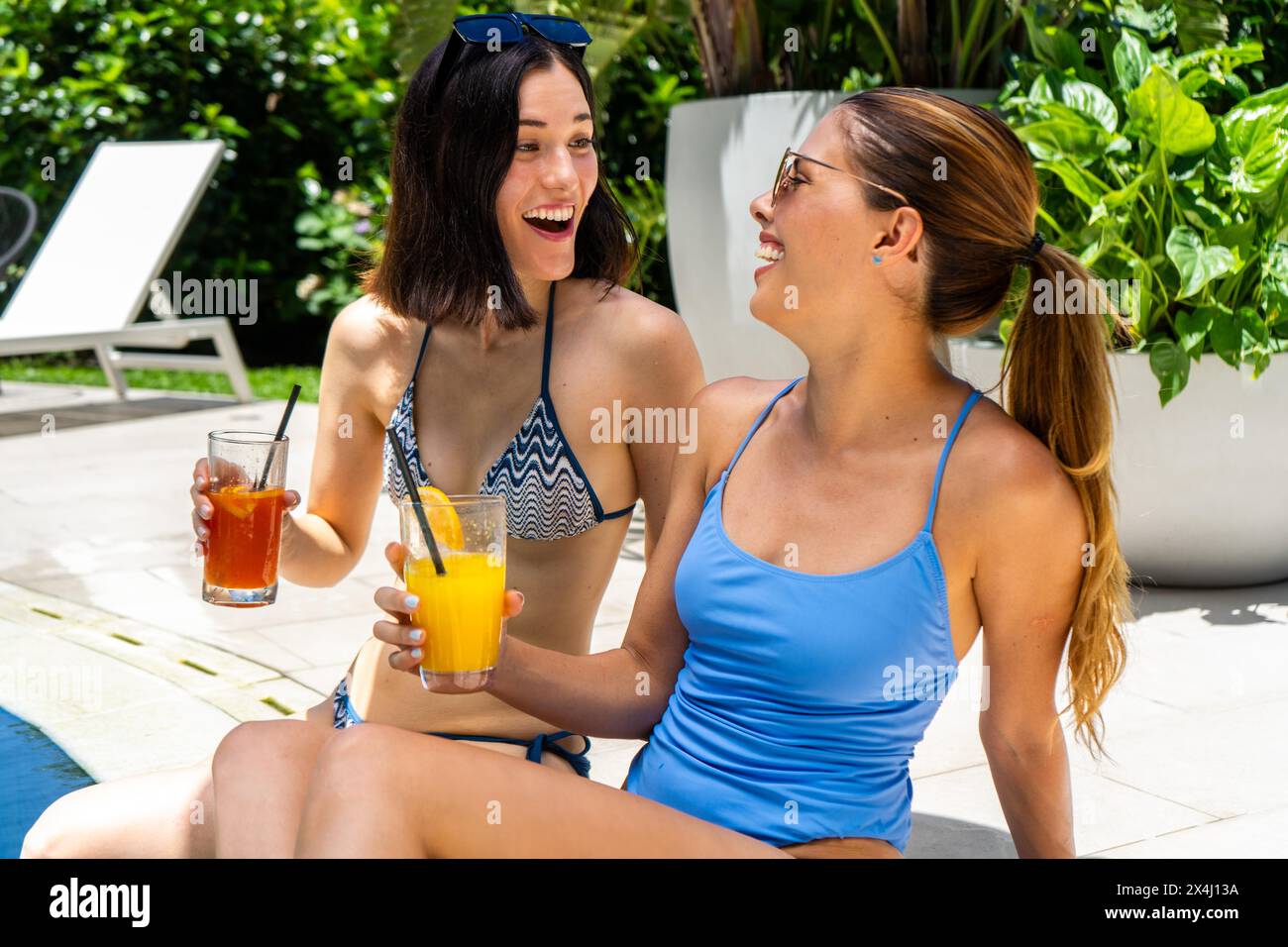 Amis heureux avec smoothies sur les mains assis côte à côte dans la piscine Banque D'Images