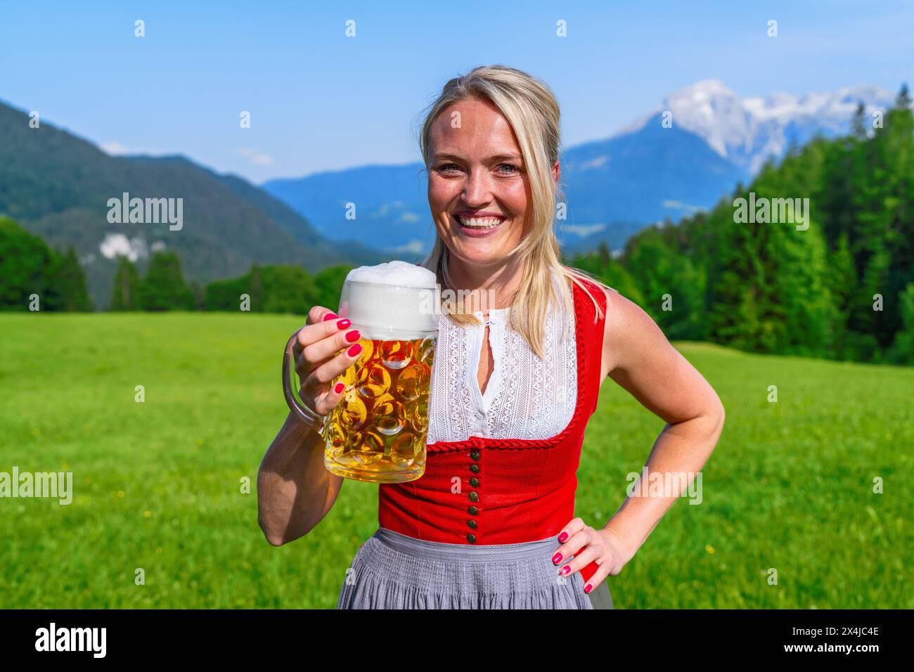 Femme blonde souriante en tracht traditionnel rouge toast avec une tasse de bière mousseuse avec des montagnes alpines en arrière-plan à l'oktoberfest bavarois Banque D'Images