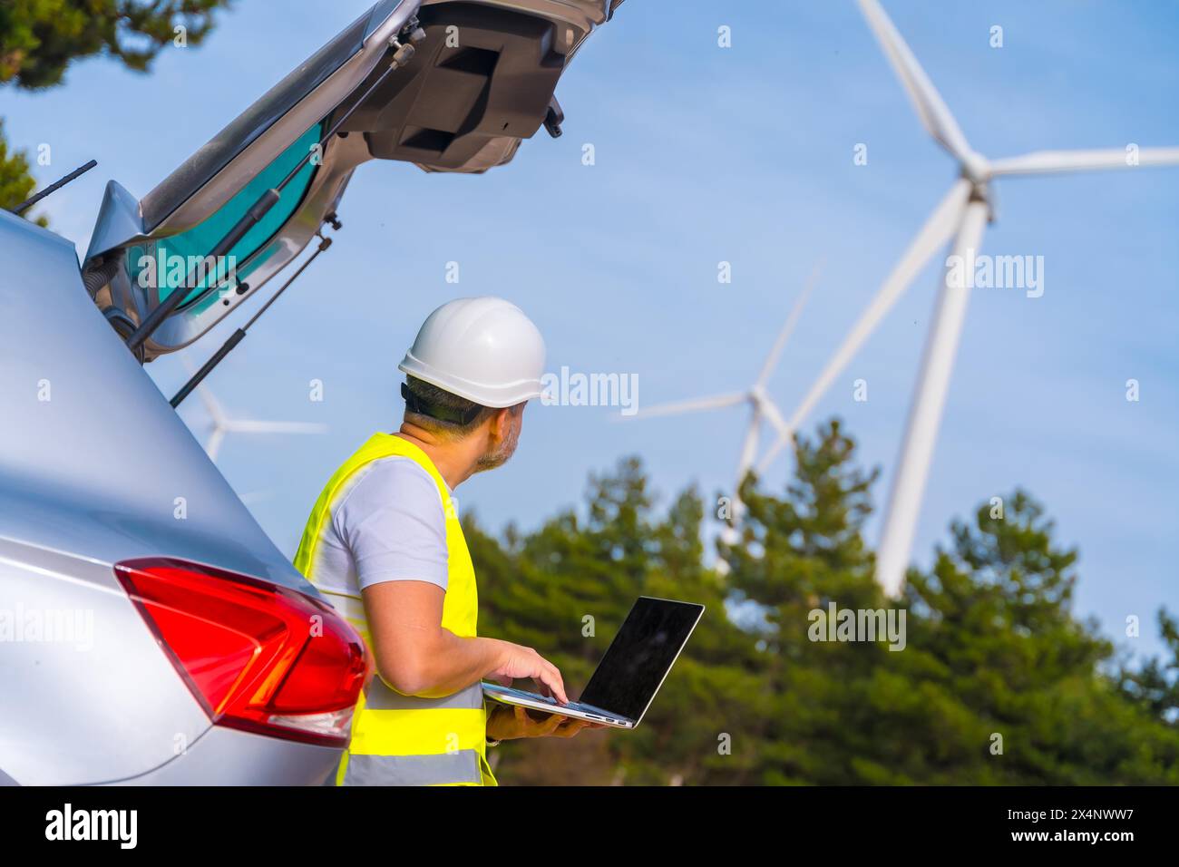 Technicien mûr masculin portant des vêtements réfléchissants et un casque rigide debout à côté de la voiture en utilisant un ordinateur portable travaillant dans un parc d'énergie éolienne avec des éoliennes Banque D'Images