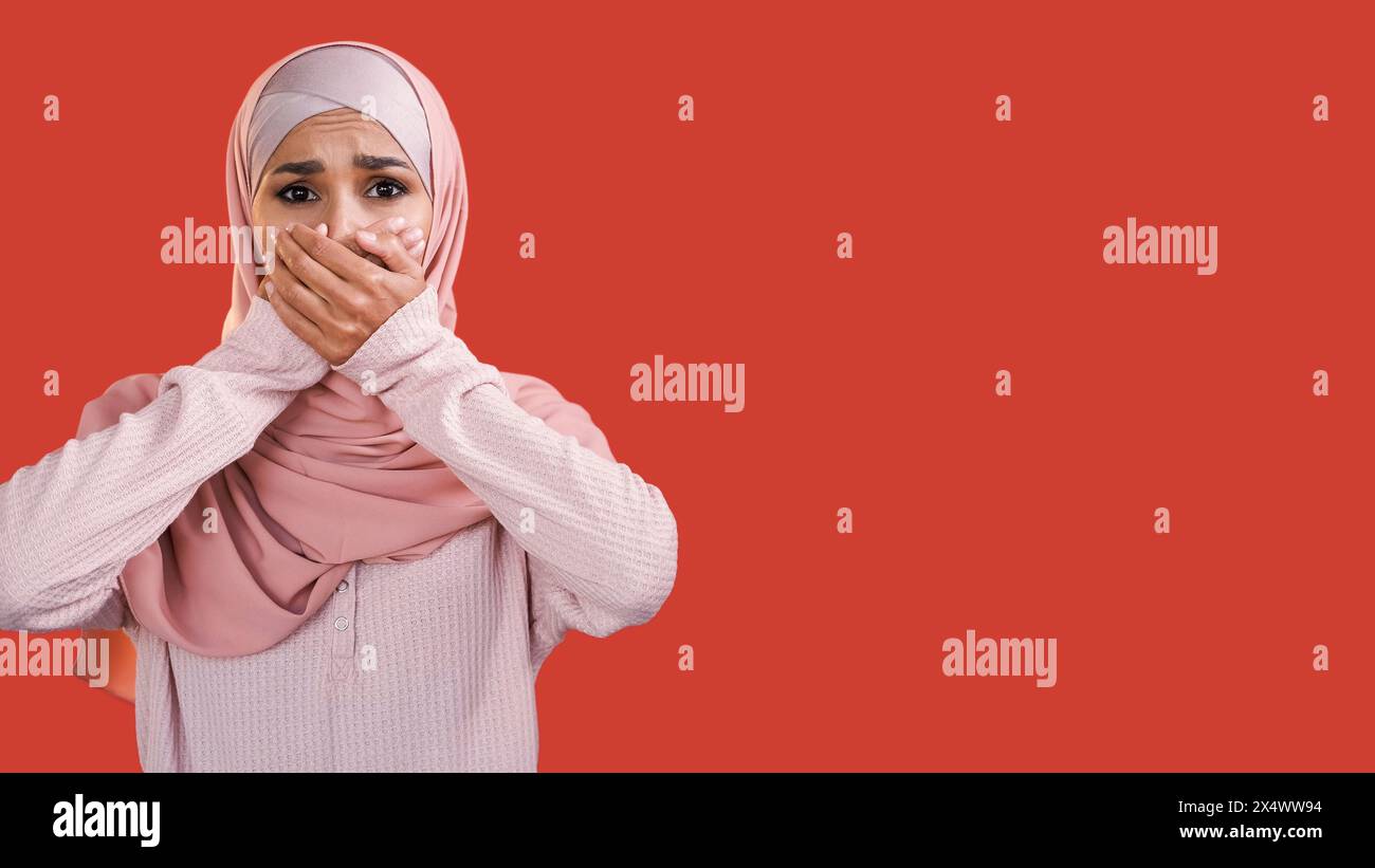 Visage effrayé. OMG panique. Des nouvelles choquantes. Femme inquiète sans voix effrayée en hijab couvrant la bouche isolée sur fond d'espace vide rouge. Banque D'Images