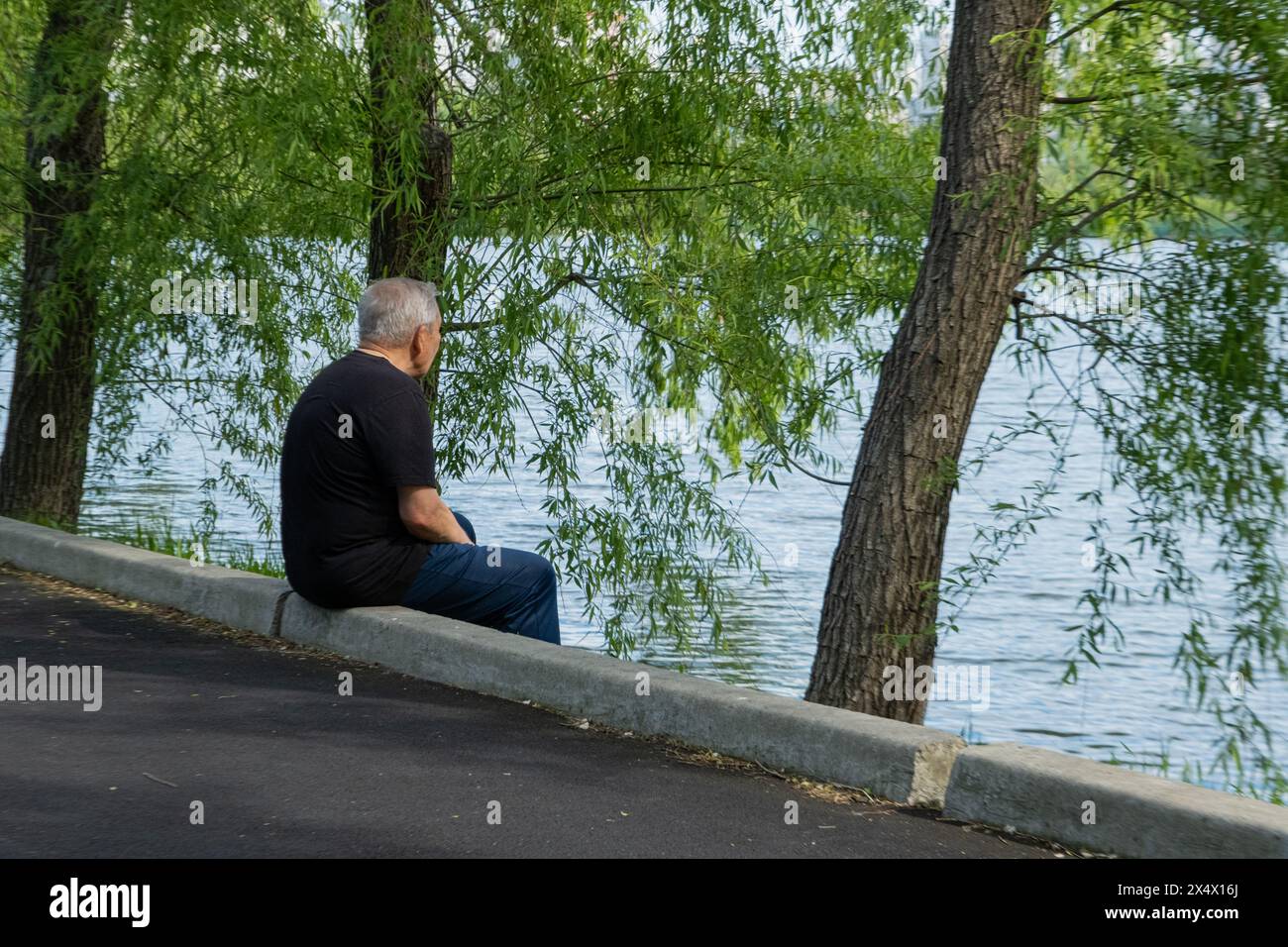 Un vieil homme en vêtements noirs est assis sur la rive au bord de l'eau et est triste de quelque chose. Vieillesse Banque D'Images