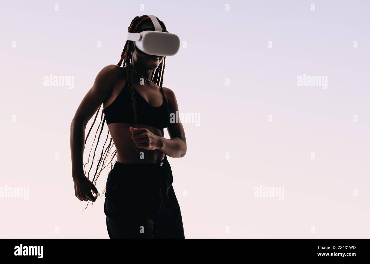 Jeune femme aux cheveux tressés danse en toute confiance dans un studio. Portant un soutien-gorge de sport, elle adopte la technologie de réalité virtuelle, en utilisant des lunettes VR. Banque D'Images