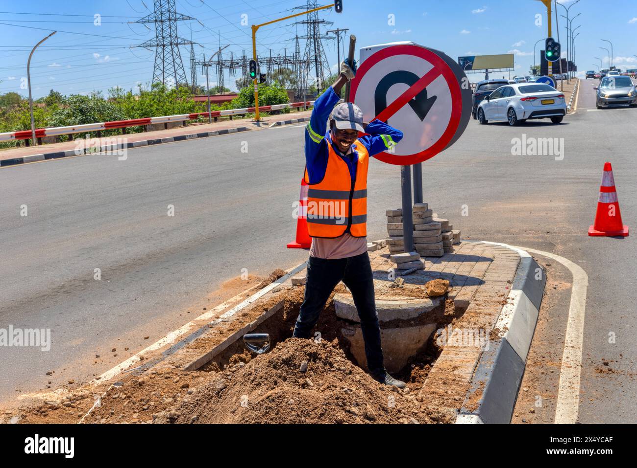 travailleur afro-américain avec une pioche creusant un fossé dans l'autoroute pour réparer le câblage sur une île à une intersection Banque D'Images