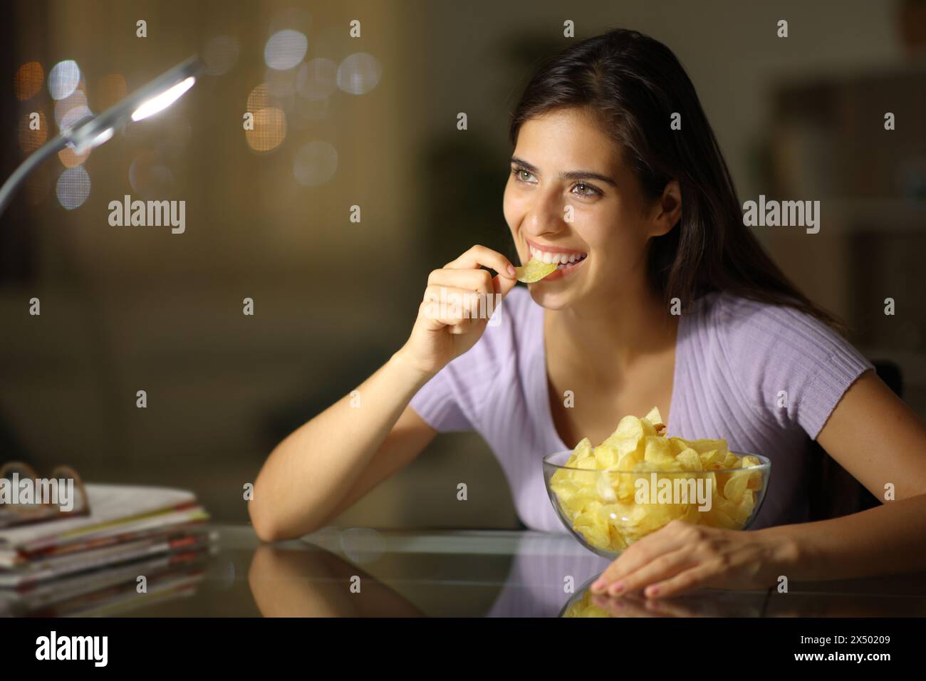 Femme heureuse dans la nuit mangeant des croustilles regardant loin à la maison Banque D'Images