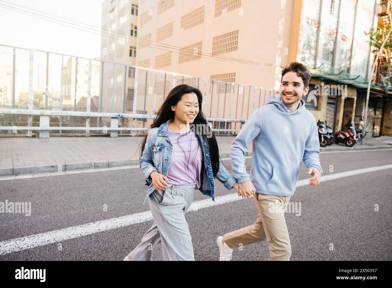 Couple interracial souriant et traversant la route se tenant par la main. Jeune homme et femme courant dans la rue de la ville. Banque D'Images