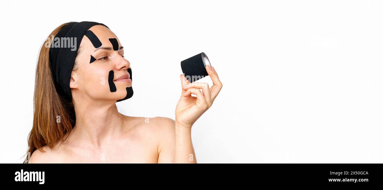 Femme modèle de beauté avec des bandes faciales kinesio sur son visage regarde le rouleau de k-tape dans sa main et souriant devant un fond blanc avec espace de copie. Banque D'Images