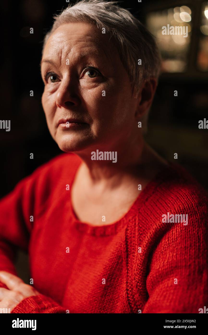 Gros plan sur la tête d'une femme âgée aux cheveux gris attrayante dans un pull rouge décontracté assis à table dans une pièce sombre par la lumière de la bougie allumée Banque D'Images