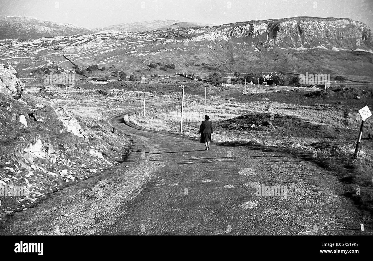 Années 1950, historique, une dame marchant sur une route dans la zone des basses terres d'Assynt, Sutherland, Highlands, Écosse. Un signe pour passer place sur la droite de l'image et la chaîne de montagnes distinctive de la zone dans la distance. Banque D'Images