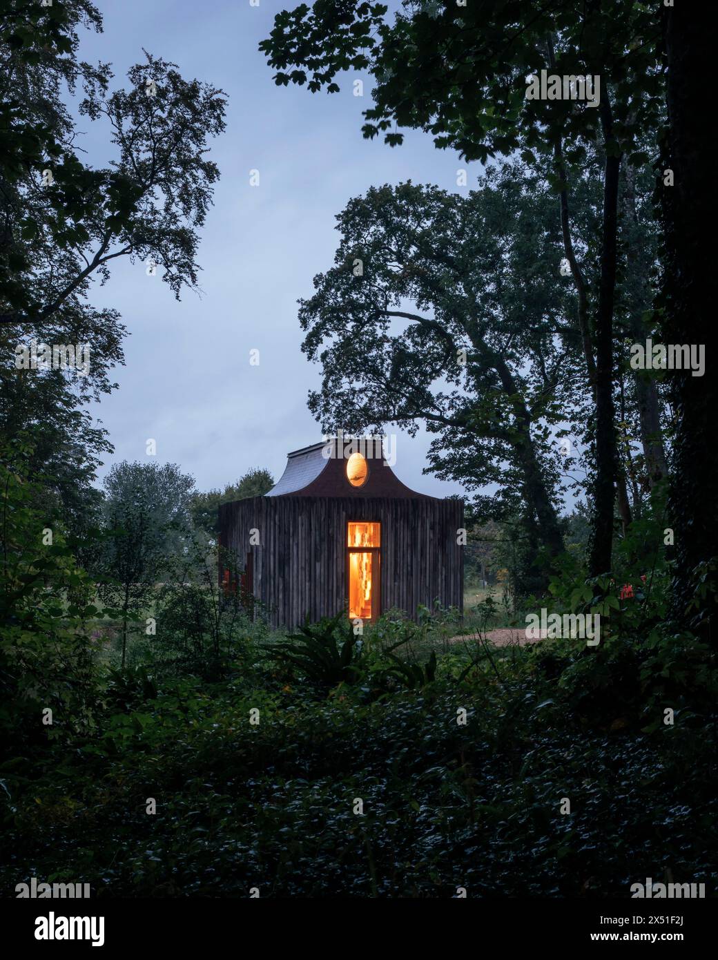 Le pavillon Apiaire vu au crépuscule, avec les arbres environnants silhouettés contre le ciel. les lumières intérieures sont dorées montrant la fenêtre de l'atrium. Le Beezantiu Banque D'Images