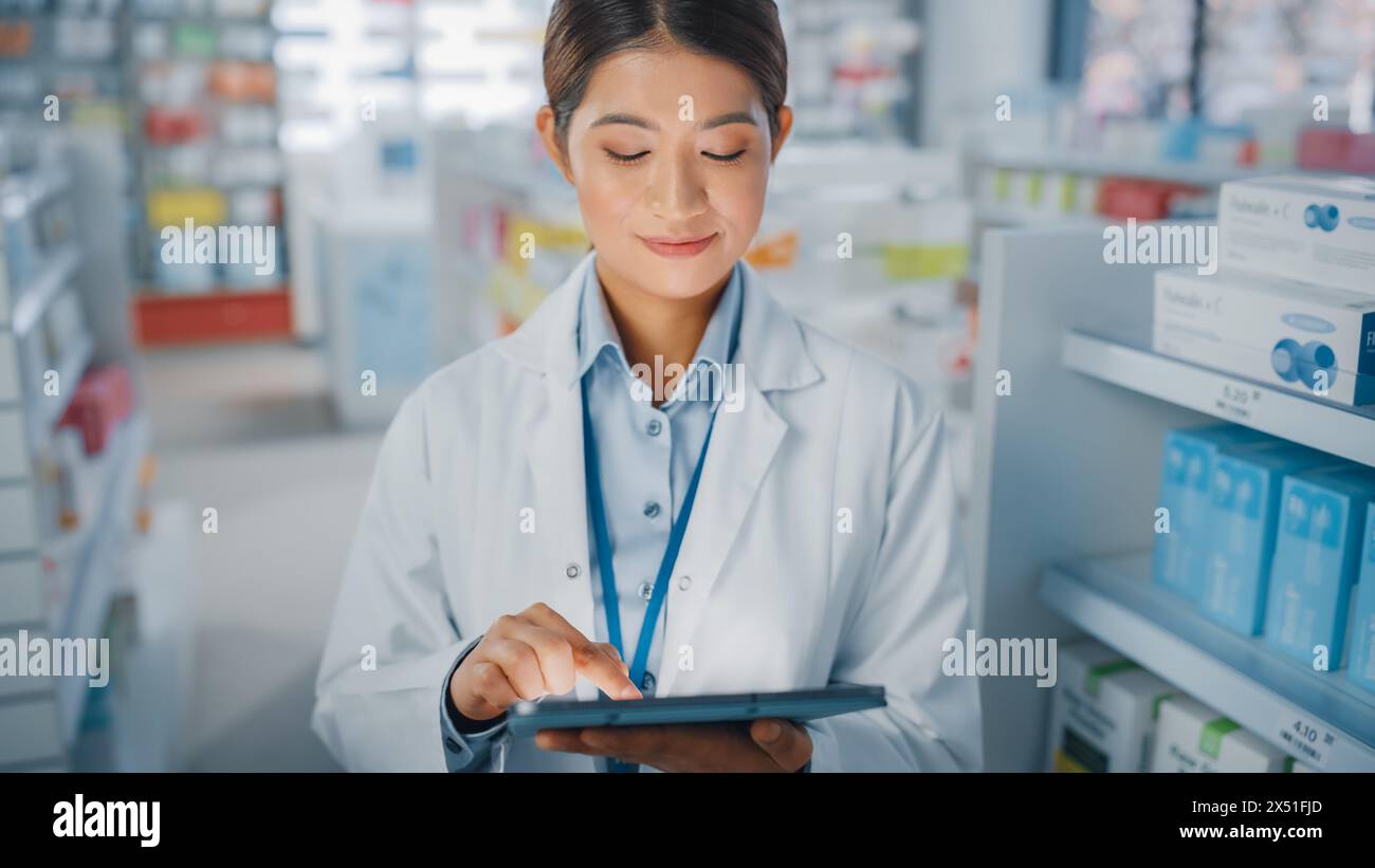 Pharmacie Drugstore : Portrait de belle pharmacienne asiatique utilise Digital Tablet Computer et sourit charmablement, derrière ses étagères pleines de paquets de médecine. Gros plan moyen Banque D'Images