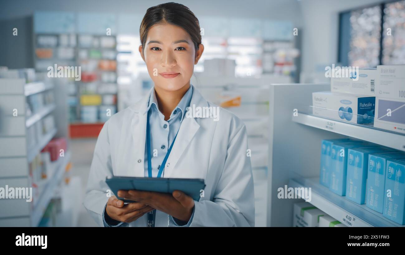 Pharmacie Drugstore : Portrait de belle pharmacienne asiatique utilise un ordinateur tablette numérique, regarde la caméra et sourit charmablement, derrière ses étagères pleines de paquets de médecine. Gros plan moyen Banque D'Images