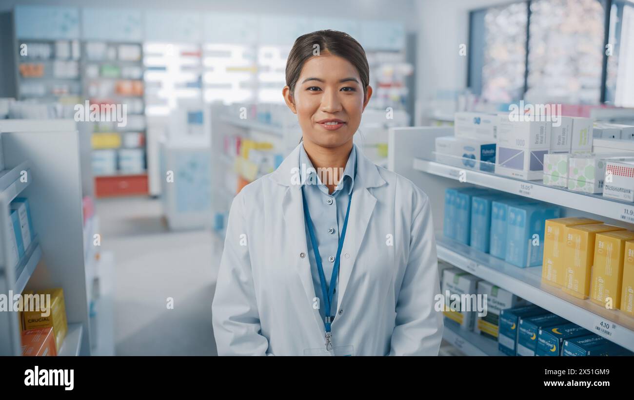 Pharmacie Drugstore : Portrait de belle pharmacienne asiatique portant un manteau blanc, regardant la caméra et souriant charmablement, derrière ses étagères pleines de paquets de médecine. Gros plan moyen Banque D'Images