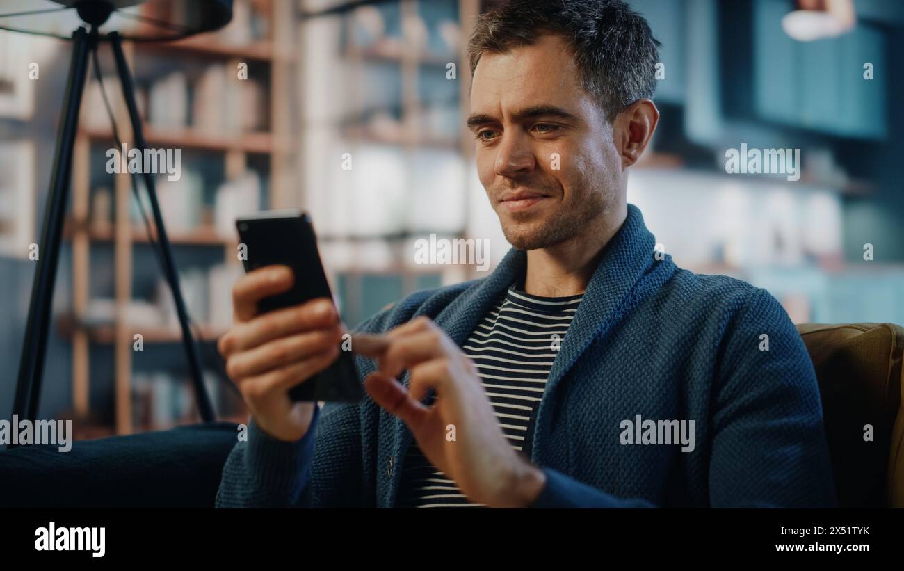 Homme caucasien excité à l'aide d'un smartphone tout en se reposant sur un canapé dans le salon élégant. Homme heureux souriant à la maison et discutant avec des collègues et des clients sur Internet. Utilisation des réseaux sociaux. Banque D'Images