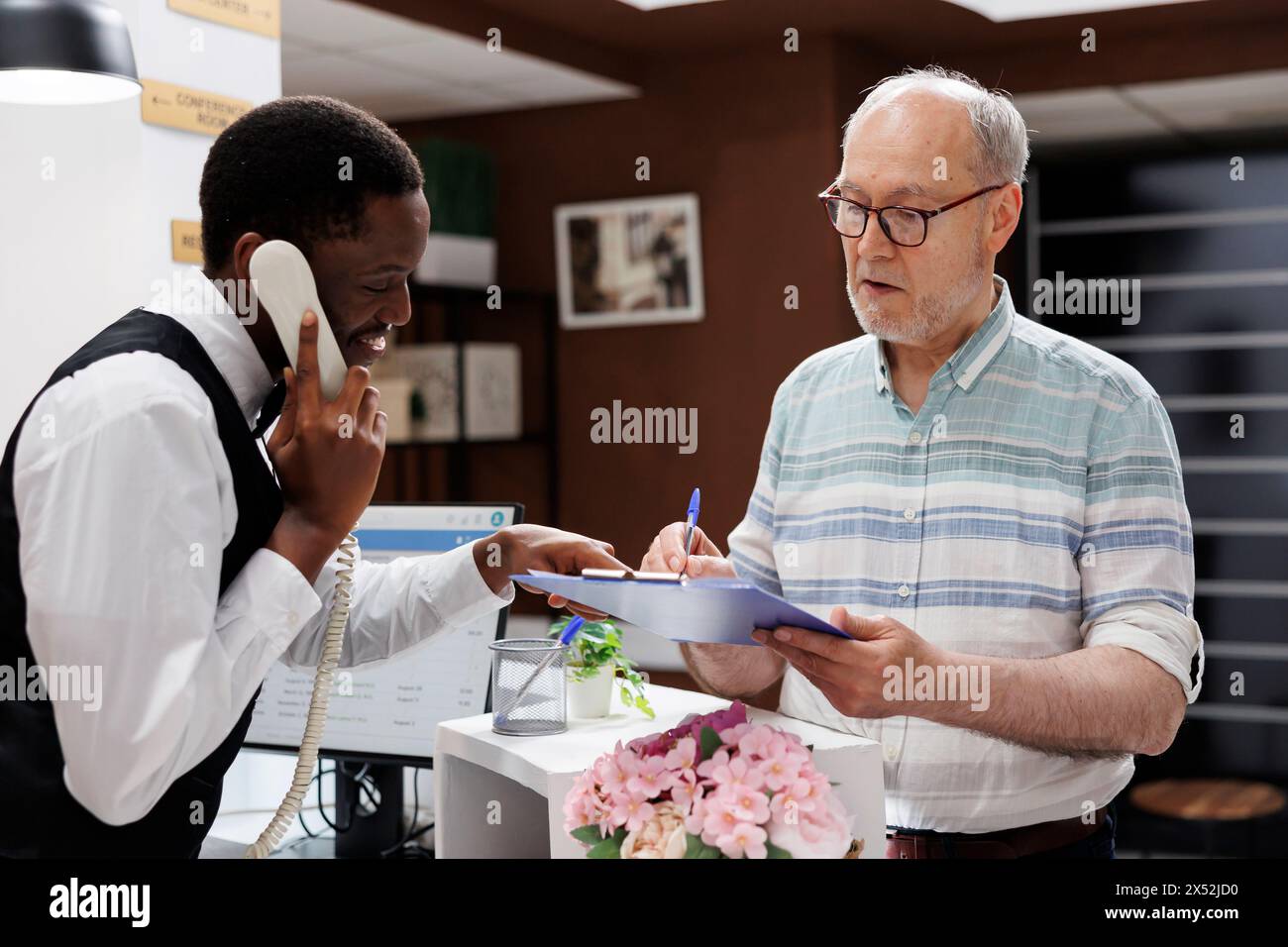 Le concierge afro-américain répond au téléphone, un vieil homme caucasien à la retraite signe les formulaires de réservation à la réception de l'hôtel. Pendant que la réceptionniste est au téléphone, le client âgé remplit les formulaires d'enregistrement. Banque D'Images