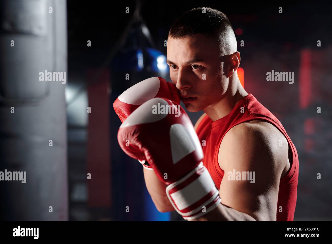 Jeune homme musclé regardant le sac de punch avant de le frapper tout en se tenant devant la caméra pendant la formation de boxe dans le gymnase Banque D'Images