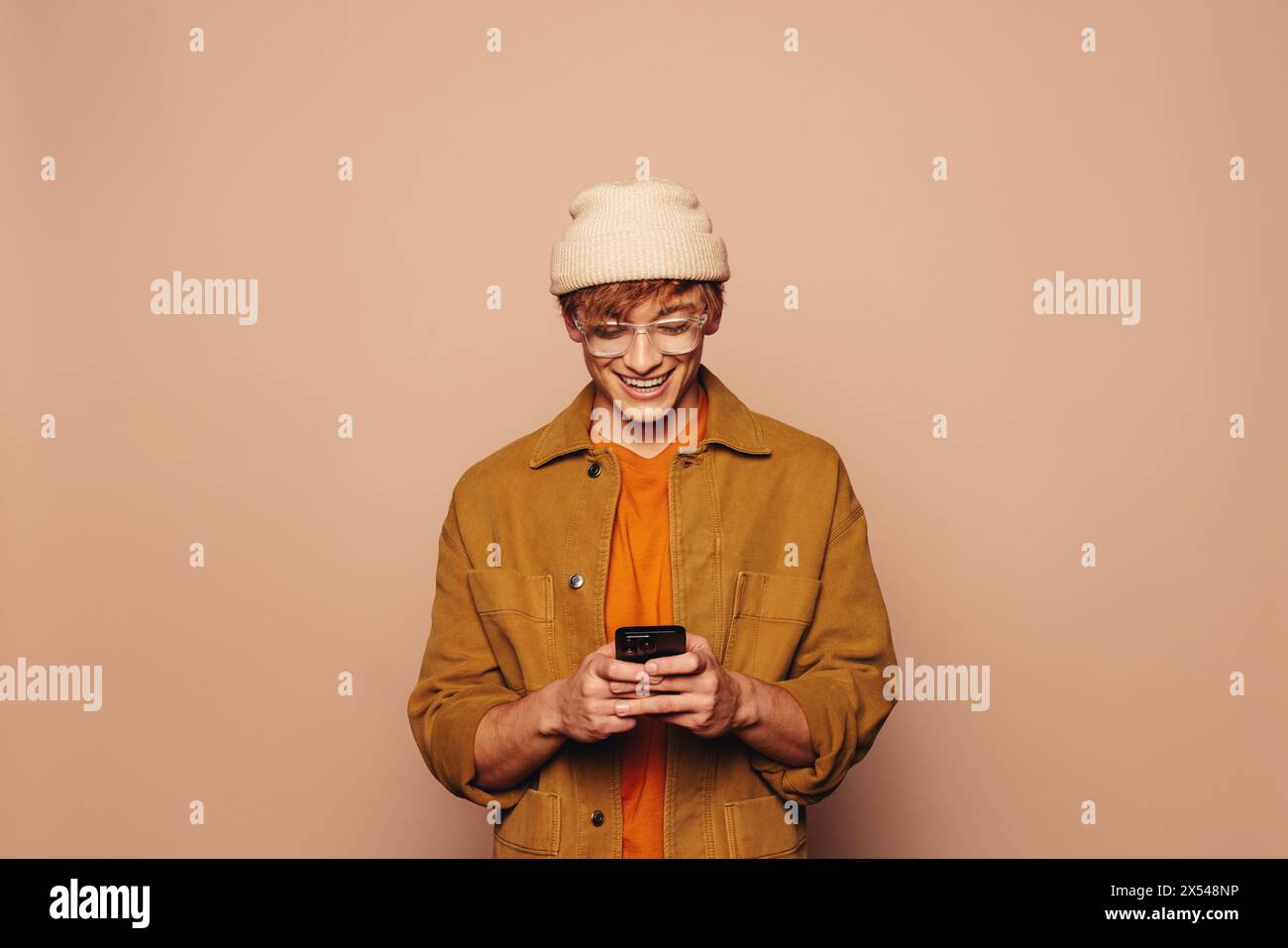 Jeune homme dans des vêtements décontractés et une veste en denim se tient sur un fond pêche. Tenant un téléphone portable, il sourit en lisant et en utilisant le social me Banque D'Images