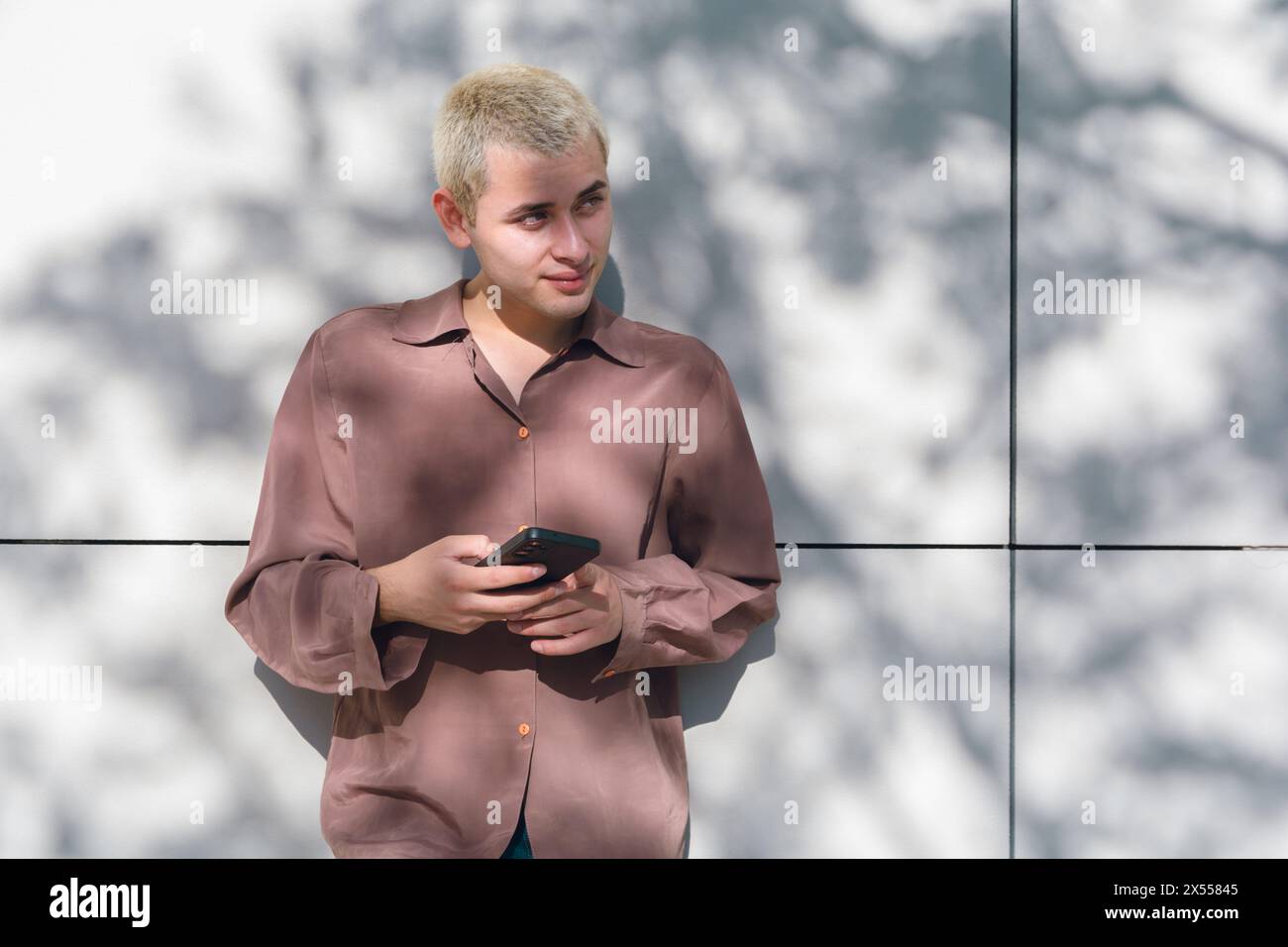 Un jeune latin aux cheveux blonds tient un téléphone portable. Il porte une chemise marron et un Jean et regarde loin Banque D'Images