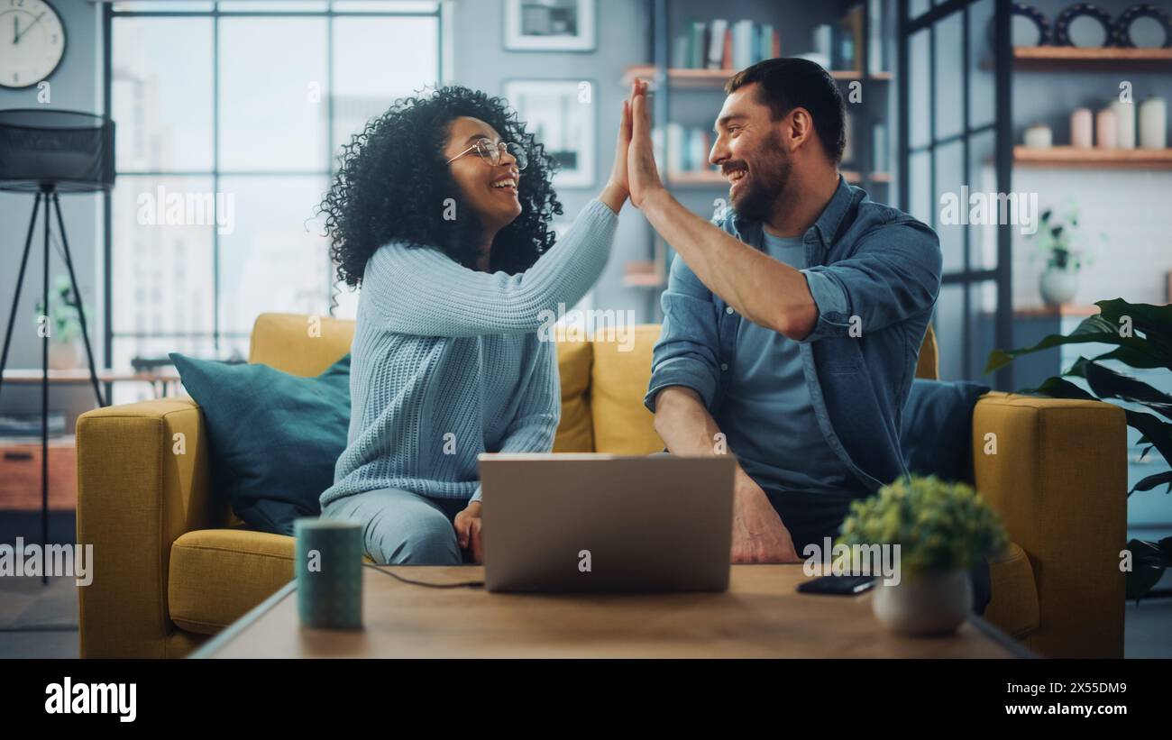 Couple multiethnique diversifié sont assis sur un canapé canapé dans le salon élégant et choisir des articles à acheter en ligne avec ordinateur portable, Give High Five. Des amis ou des collègues discutent de projets de travail. Banque D'Images