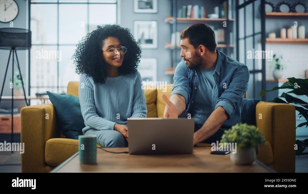Couple multiethnique diversifié sont assis sur un canapé canapé dans le salon élégant et choisir des articles à acheter en ligne avec un ordinateur portable. Des amis ou des collègues discutent de projets de travail. Banque D'Images