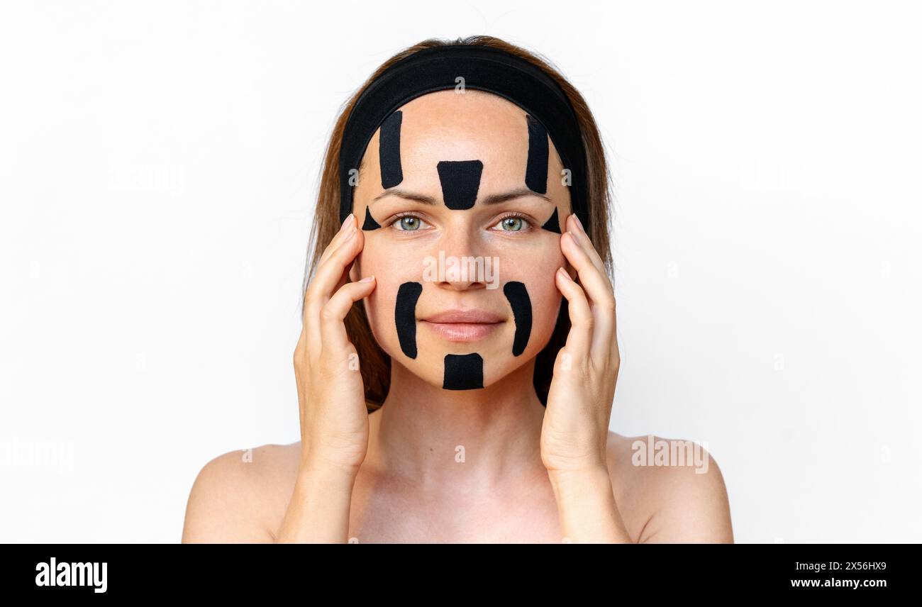 Rajeunissement facial et thérapie alternative anti-âge. Modèle de beauté femme brune isolé appliquant des bandes faciales kinesio sur son visage devant whi Banque D'Images