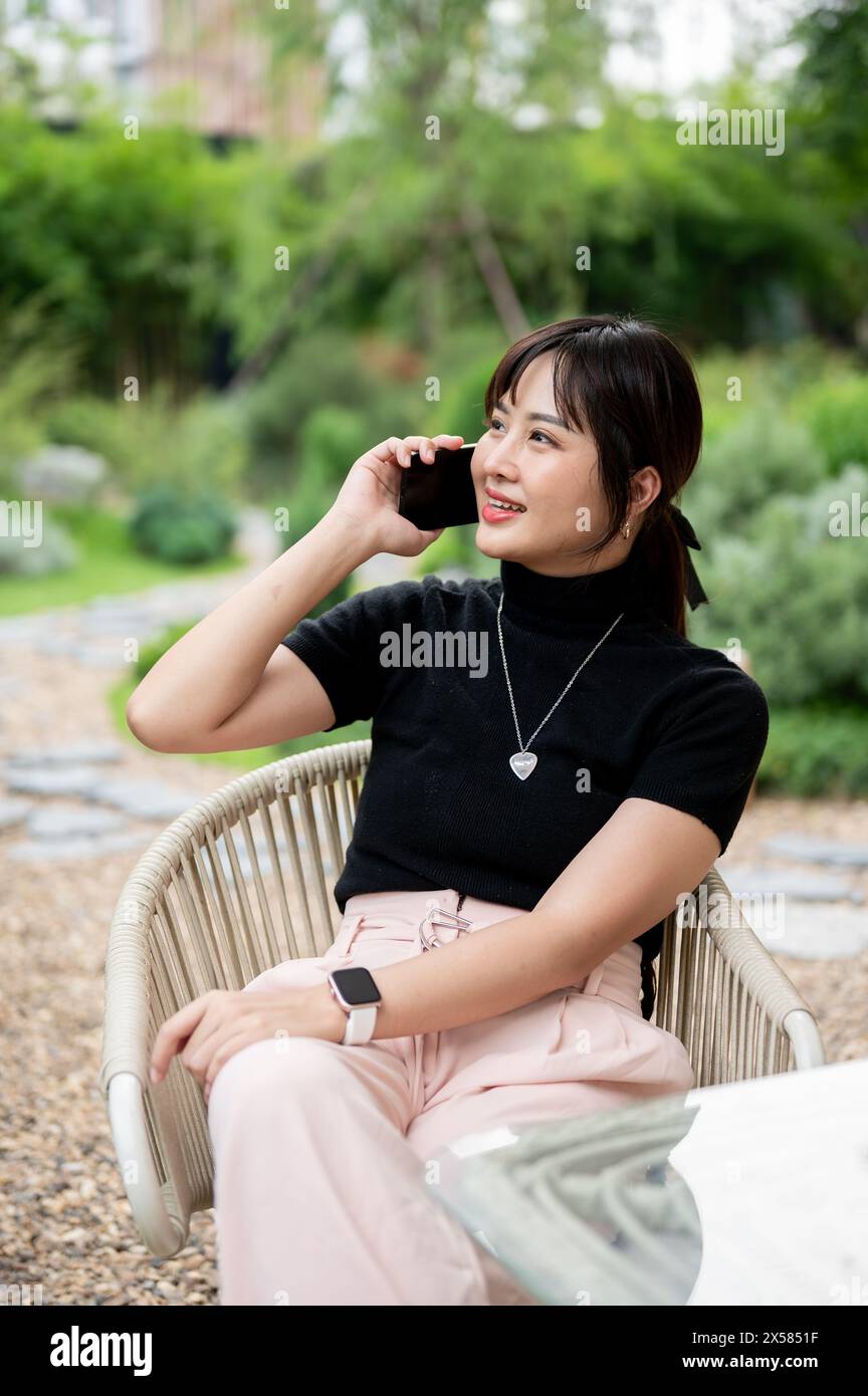 Une belle femme asiatique positive dans des vêtements décontractés se relaxant dans un jardin verdoyant ou arrière-cour, assise à une table et parlant au téléphone. des gens, des vies Banque D'Images