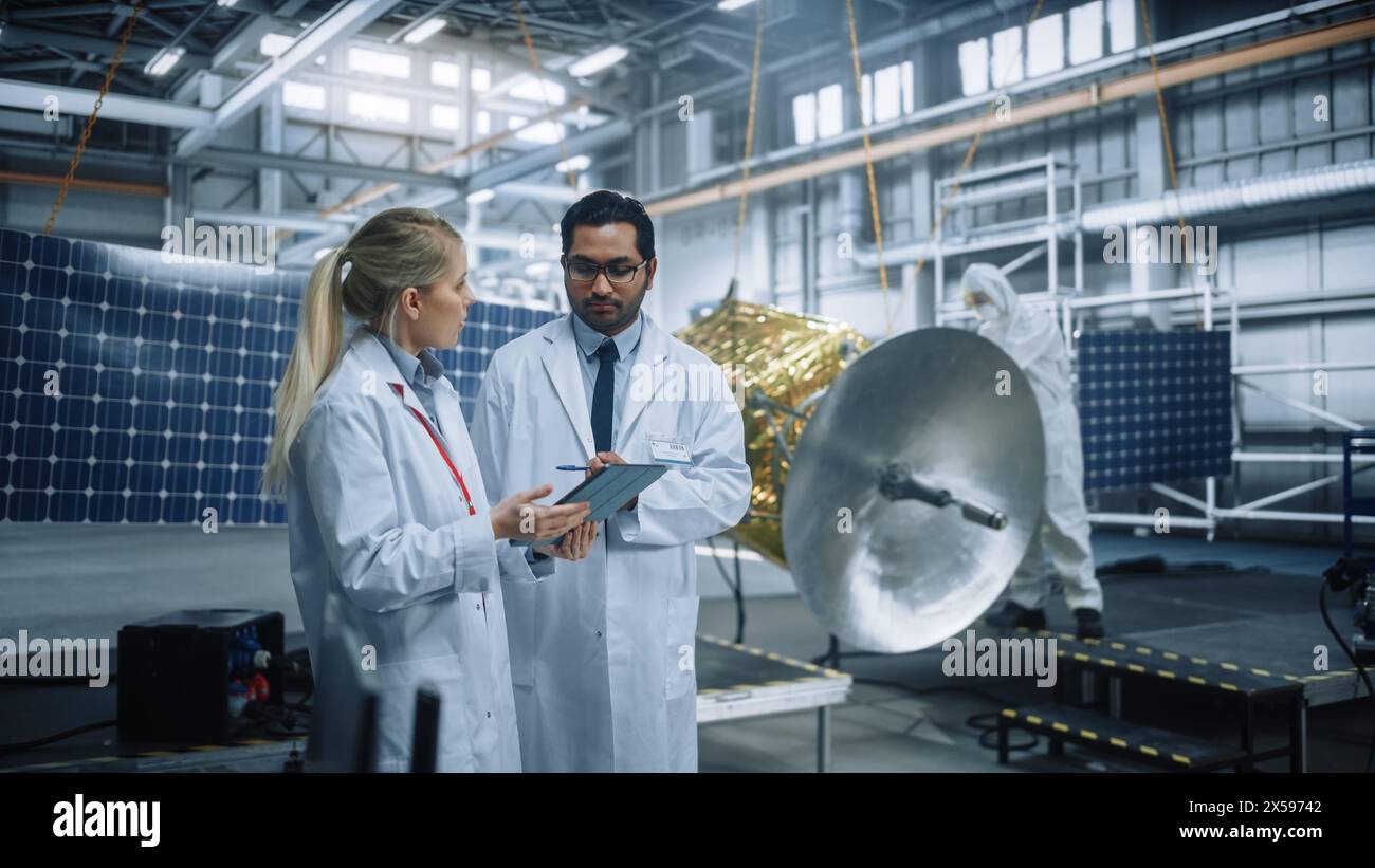 Deux ingénieurs parlent, utilisent un ordinateur tablette tout en travaillant sur la construction de satellites. Installation de fabrication de l'Agence aérospatiale : les scientifiques construisent et assemblent des engins spatiaux pour la mission d'exploration spatiale Banque D'Images