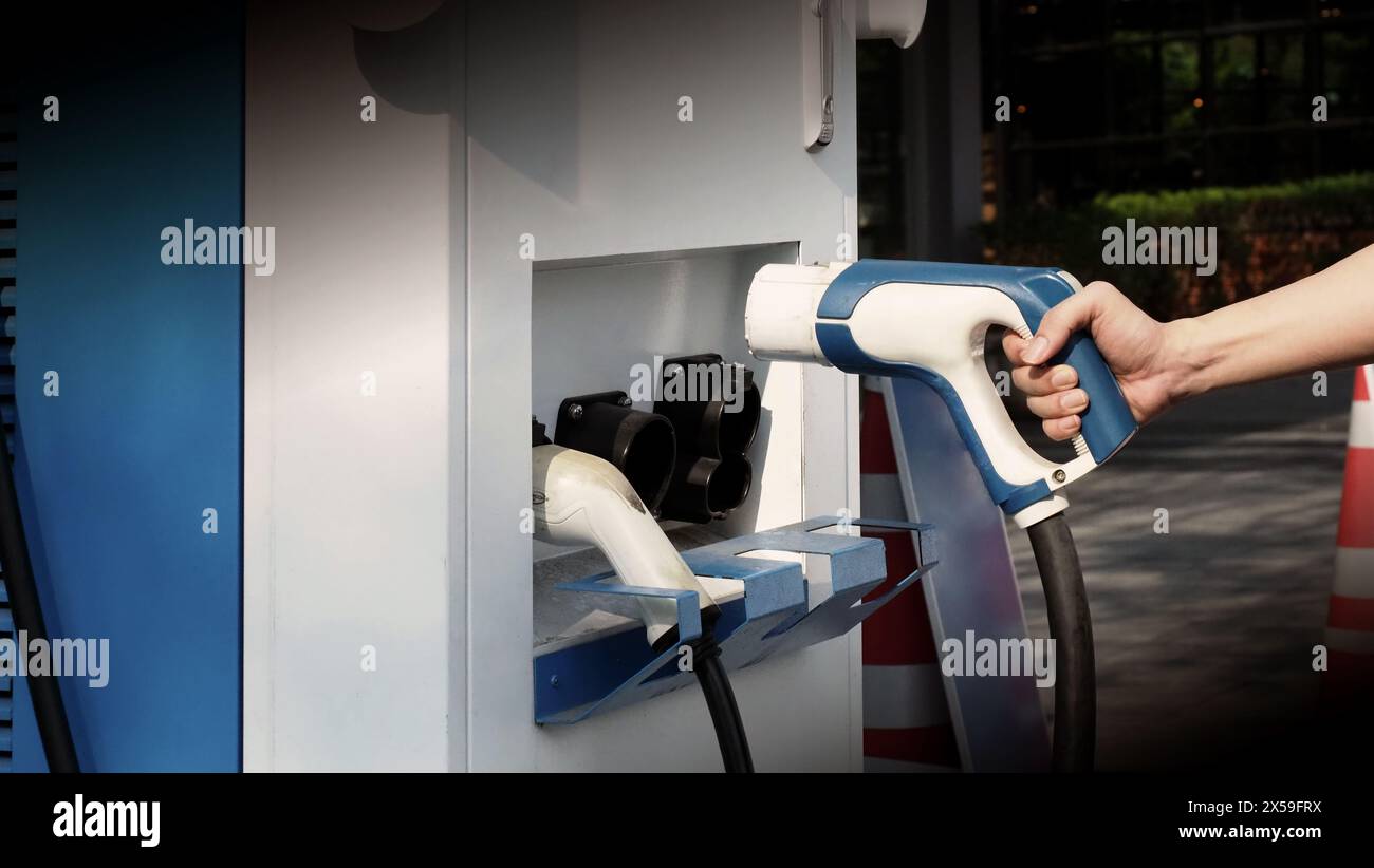 Le conducteur de la voiture électrique tient la prise du chargeur de batteries. Main tenant la prise de charge EV devant l'appareil photo avec arrière-plan flou de l'extérieur. Durable e Banque D'Images