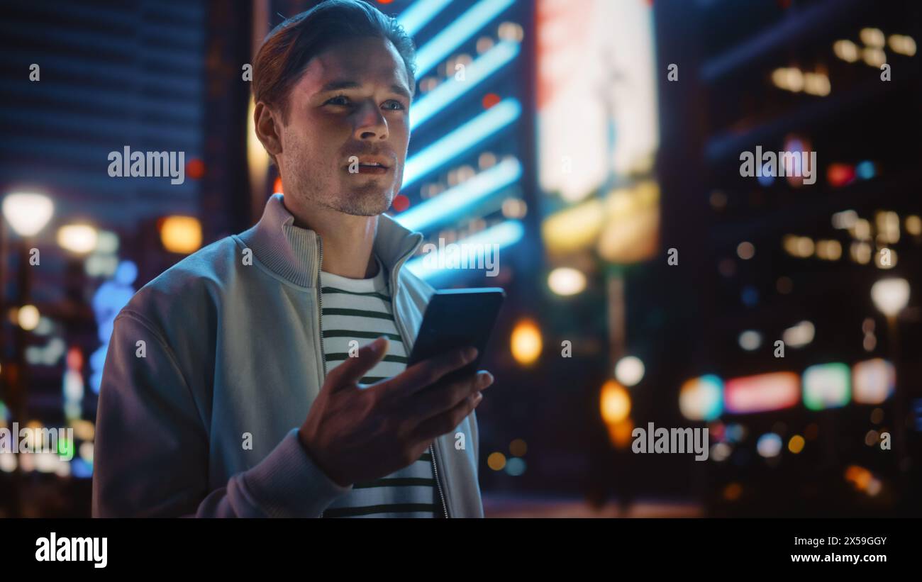 Portrait de l'homme à l'aide d'un smartphone marchant dans la rue de la ville de nuit pleine de néon lumière. Homme élégant souriant à l'aide de téléphone portable, médias sociaux, achats en ligne, SMS sur Dating App. Banque D'Images