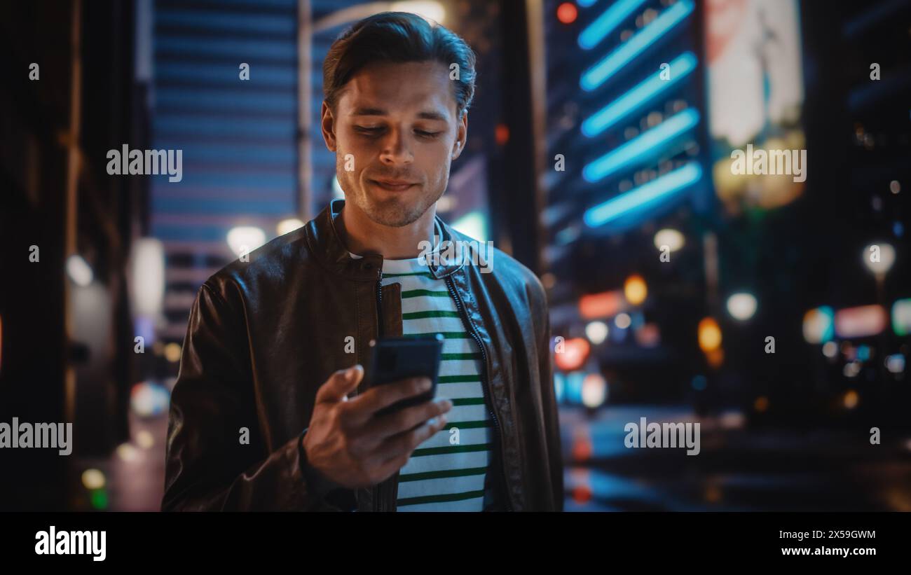 Homme utilisant un smartphone marchant dans la rue de nuit de la ville pleine de néon lumière. Homme élégant souriant à l'aide de téléphone portable, médias sociaux, achats en ligne, SMS sur Dating App. Banque D'Images