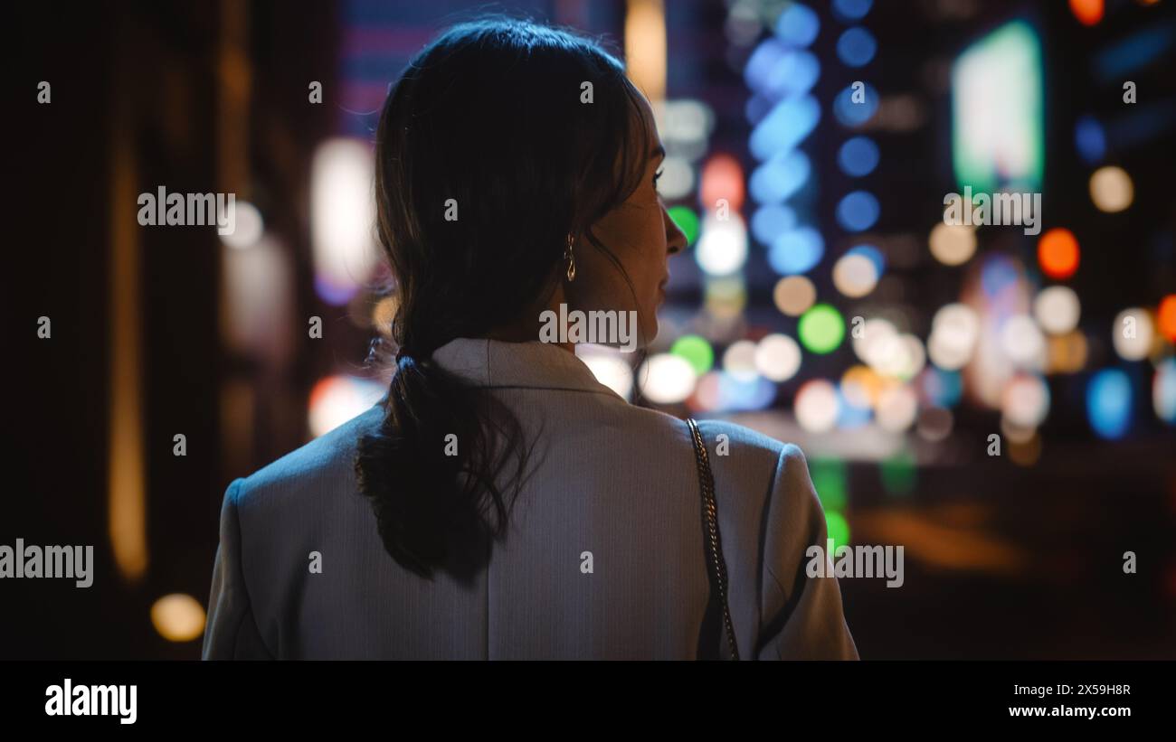 Suite à la photo de belle femme marchant dans une rue moderne de la ville avec des néons lumières la nuit, regardant à droite. Femme confiante marchant dans la zone urbaine. Vue arrière. Banque D'Images