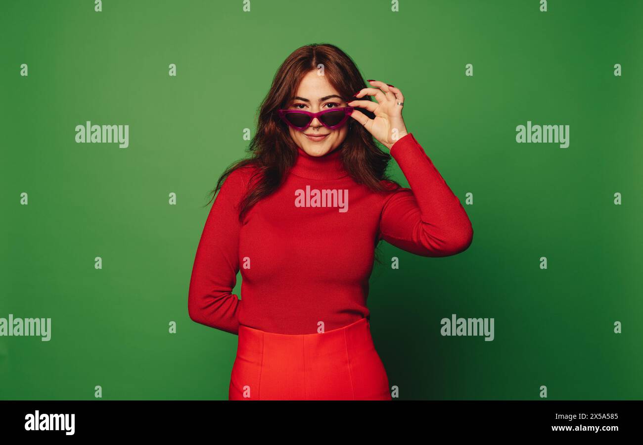 Élégamment tendance, une femme se tient dans un studio parmi les couleurs vives de vert et de rouge. Jetant un œil ludique, elle porte des vêtements décontractés, du verre à la mode Banque D'Images