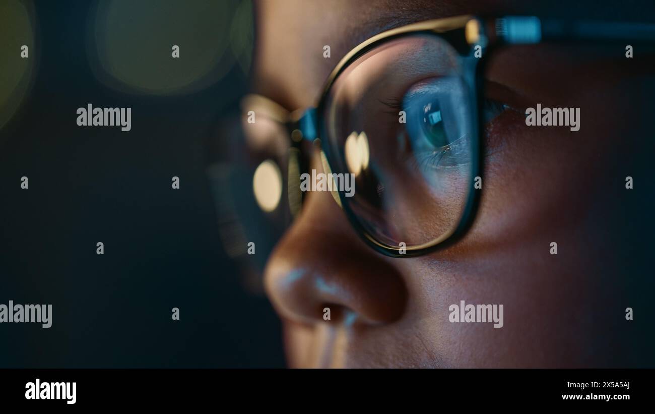 Gros plan Portrait d'une jolie jeune adolescente multiethnique fille noire dans une chambre confortable sombre à la maison. Jeune femme porte des lunettes qui reflètent les devoirs scolaires de son écran d'ordinateur portable en eux. Banque D'Images