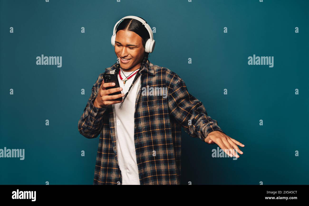 Homme heureux et occasionnel avec des cheveux tressés se tient dans un studio. Il écoute de la musique sur son téléphone portable, avec un fond bleu. Banque D'Images