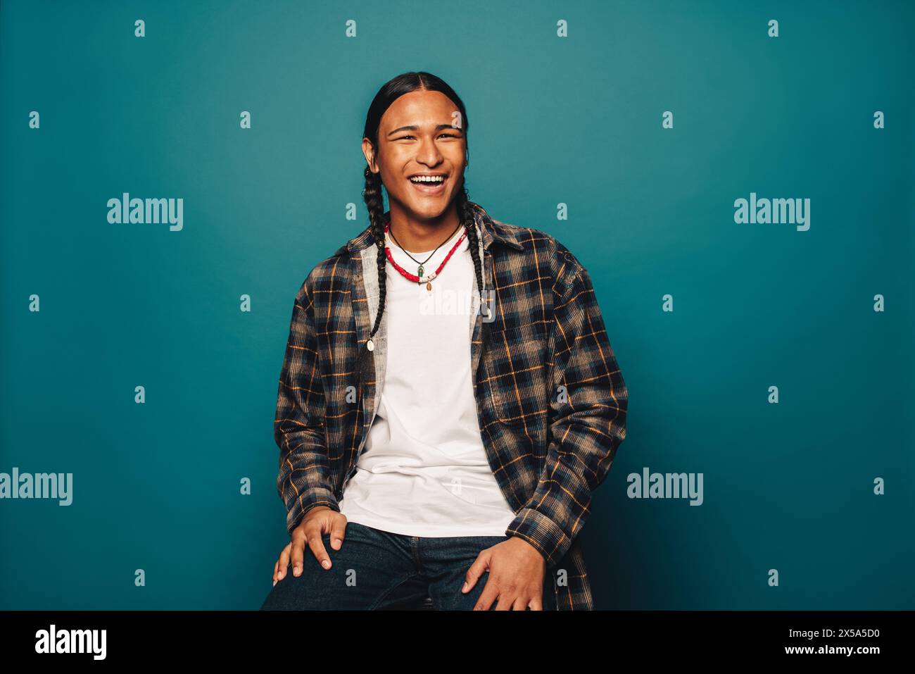 Confiant, heureux homme autochtone avec des cheveux tressés et des bijoux ethniques pose dans un studio portant des vêtements décontractés et un fond bleu. Banque D'Images