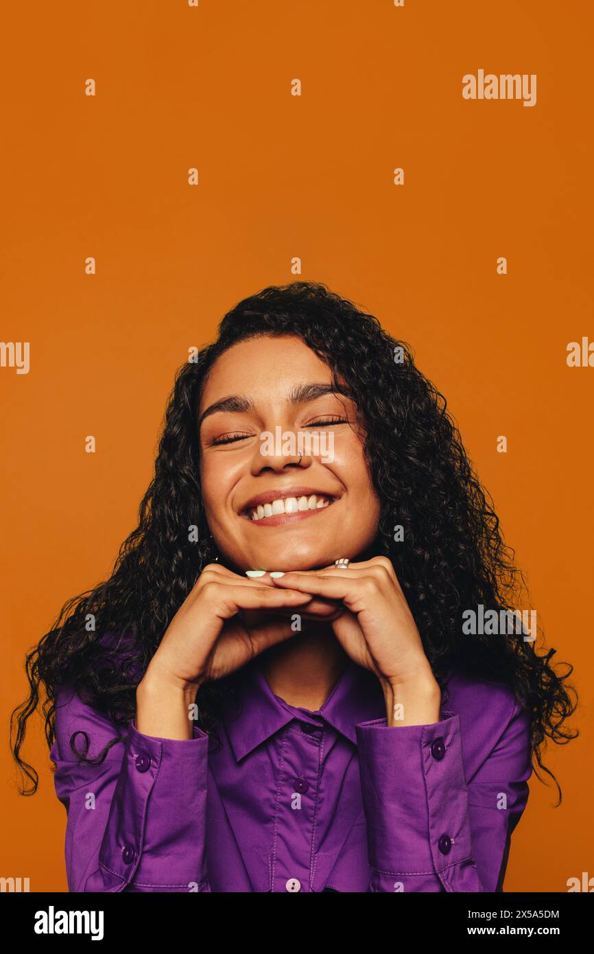 Femme joyeuse avec des cheveux bouclés et un sourire vibrant dans des vêtements décontractés. Sur un fond orange, son visage expressif et son expression joyeuse brillent. Banque D'Images