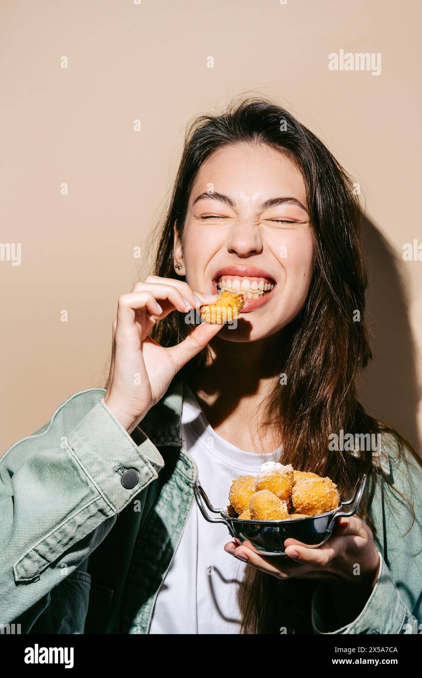 Une jeune femme joyeuse mord dans une boule de fromage frite croustillante, tenant un bol rempli de collations plus délicieuses. Banque D'Images