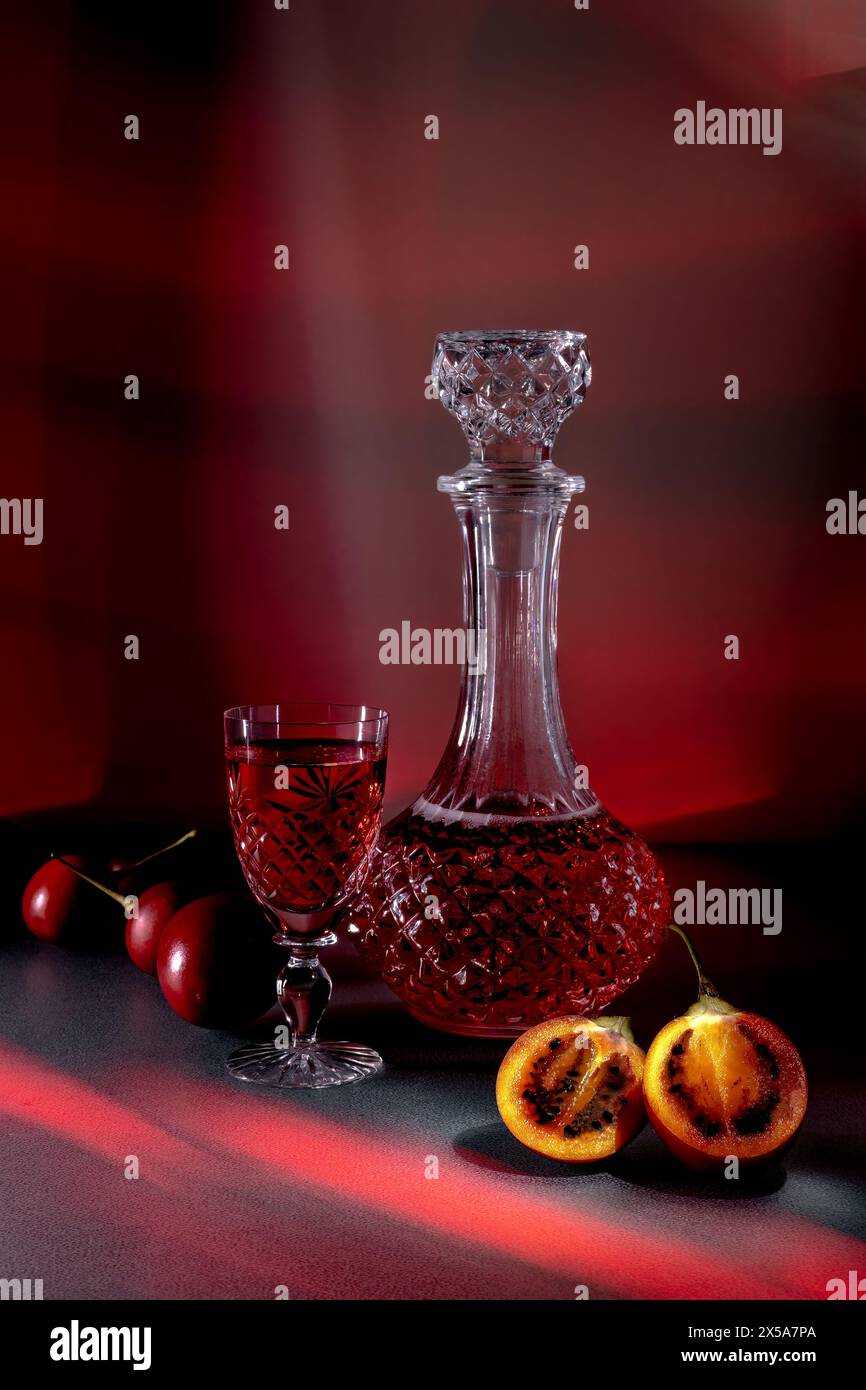 Un affichage sophistiqué avec une carafe en cristal remplie de liquide rouge, accompagné d'un verre assorti, de cerises fraîches et de tamarillo coupé en deux Banque D'Images