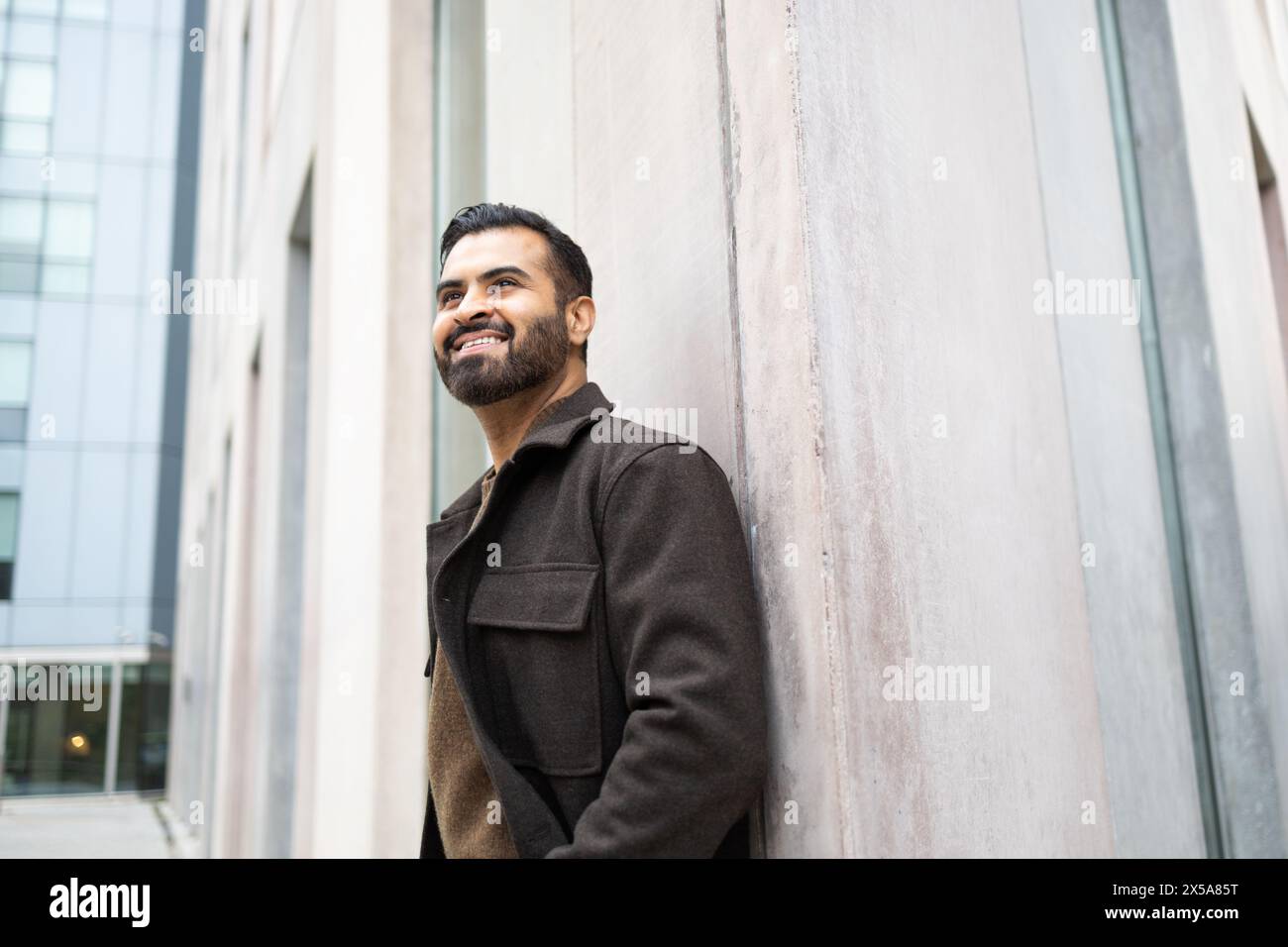 Un homme content avec une barbe dans un manteau brun appuyé contre un mur de bâtiment urbain, regardant vers le haut et souriant Banque D'Images