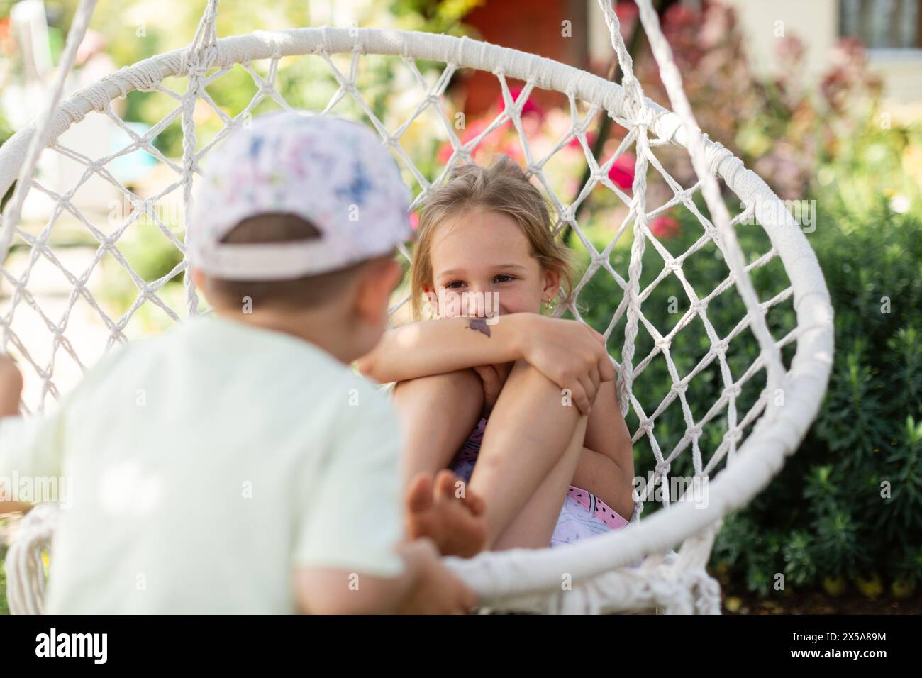 Deux jeunes frères et sœurs s'engagent dans un jeu joyeux sur une balançoire blanche dans un jardin verdoyant, se prélassent dans la chaleur d'une journée ensoleillée et partagent un moment de happ Banque D'Images