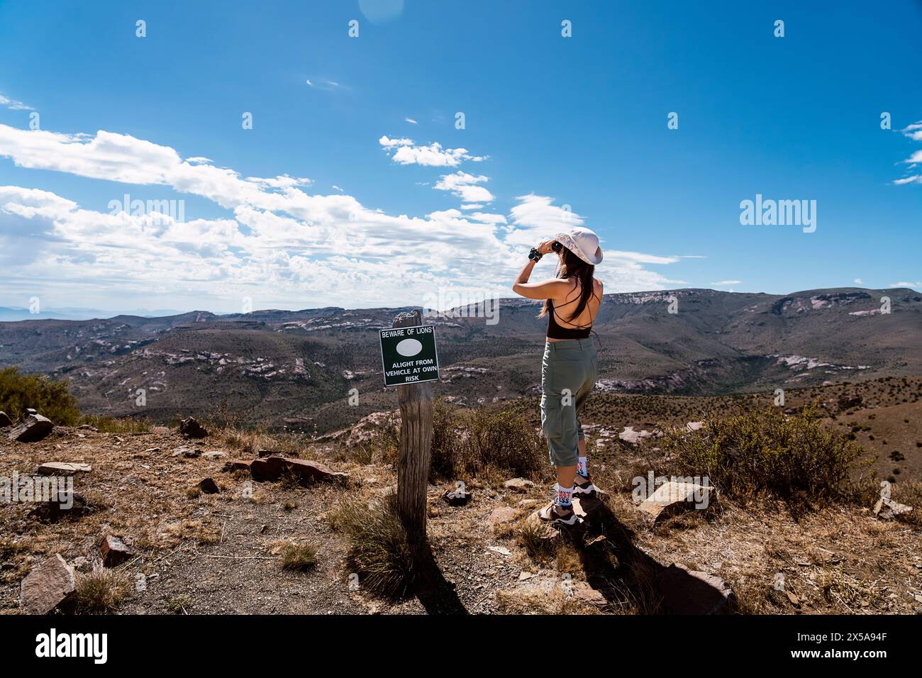 Vue arrière d'une femme randonneuse utilisant des jumelles à côté d'un panneau d'avertissement dans un paysage de montagne pittoresque sous un ciel bleu. Banque D'Images
