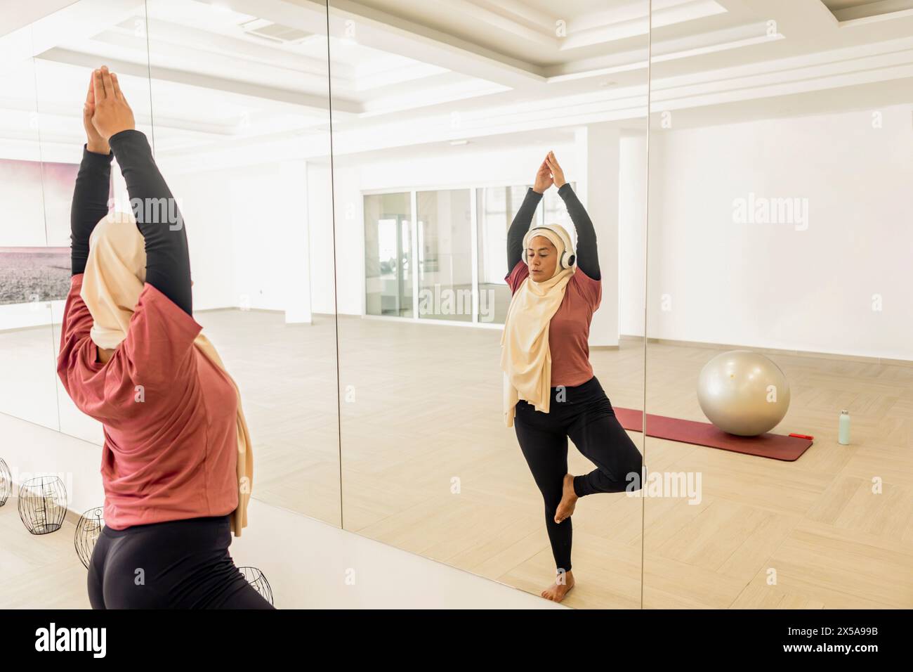 Une femme musulmane en hijab exécute la pose Vrksasana, ou pose arbre, dans un studio de yoga, réfléchissant sur un mur en miroir. Banque D'Images