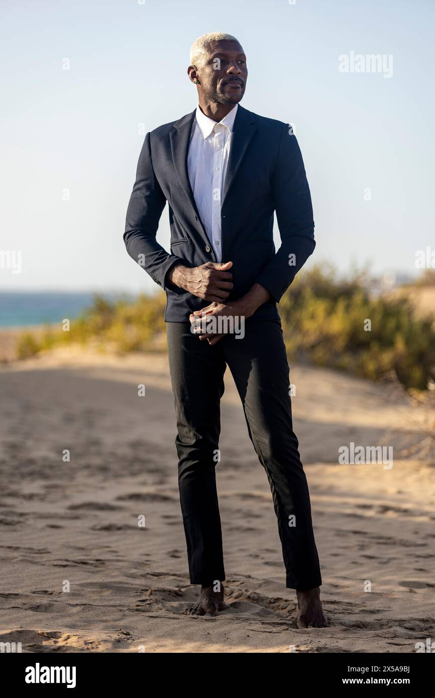 Un mannequin afro-américain pose en toute confiance sur une plage de sable, vêtu d'un élégant costume noir et d'une chemise blanche Banque D'Images