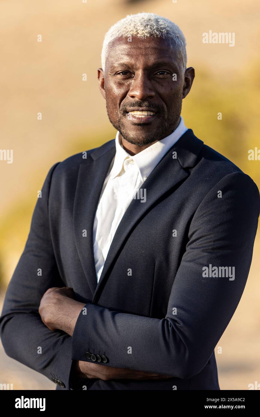 Un homme afro-américain digne aux cheveux blancs distinctifs pose en toute confiance à l'extérieur dans un costume pointu, exsudant le professionnalisme Banque D'Images