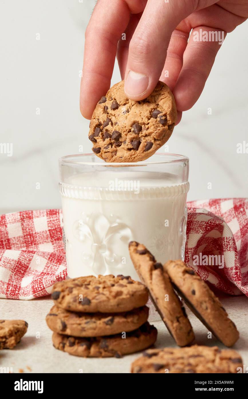 Une main est capturée en plaçant un biscuit aux pépites de chocolat sur un grand verre de lait, avec une pile de biscuits et une serviette à carreaux à côté Banque D'Images