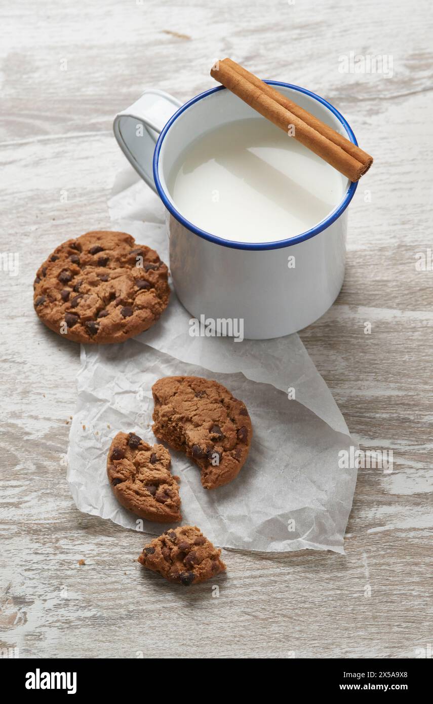 Une tasse rustique de lait garnie d'un bâton de cannelle à côté des biscuits aux pépites de chocolat fraîchement cuits sur une table en bois altérée Banque D'Images