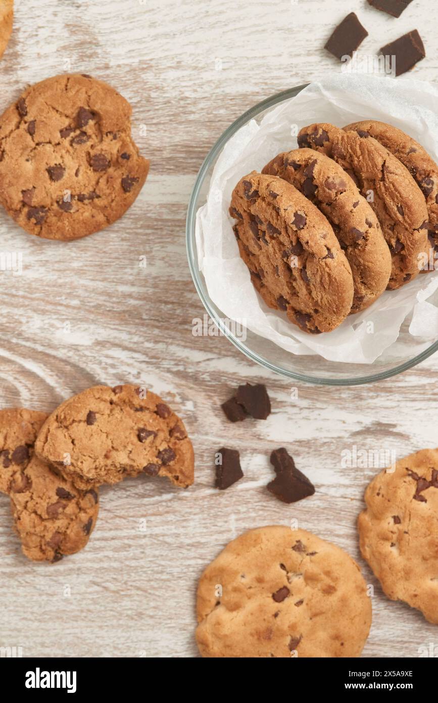 Biscuits aux pépites de chocolat dans un bol en verre et dispersés sur une table en bois rustique, photographiés d'en haut Banque D'Images