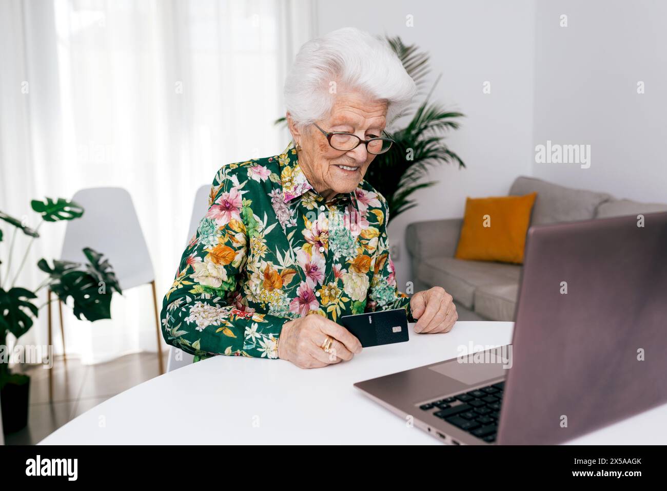Une femme âgée dans une chemise florale sourit tout en tenant une carte de crédit devant un ordinateur portable, s'engageant dans des achats en ligne dans le confort de sa maison Banque D'Images